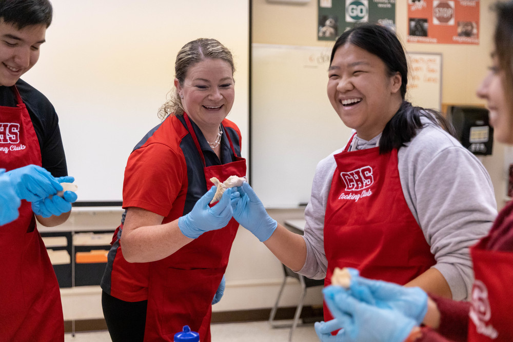 Teacher and two students in red aprons, blue gloves, "cheers" with pot stickers