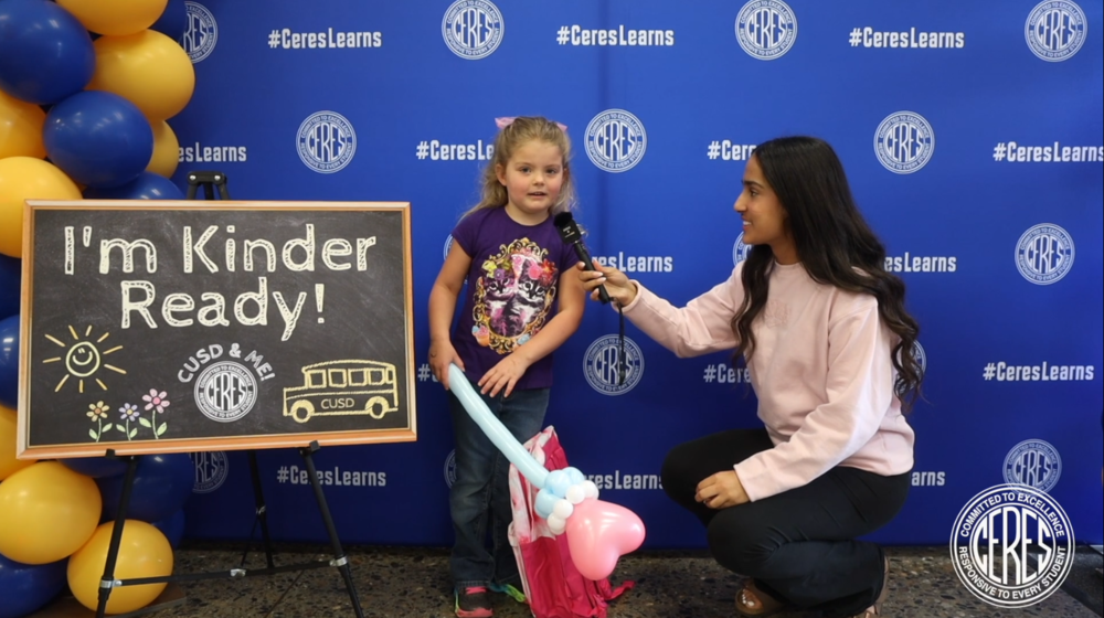 High school student in pink sweater interviews little girl next to "I'm Kinder Ready" sign