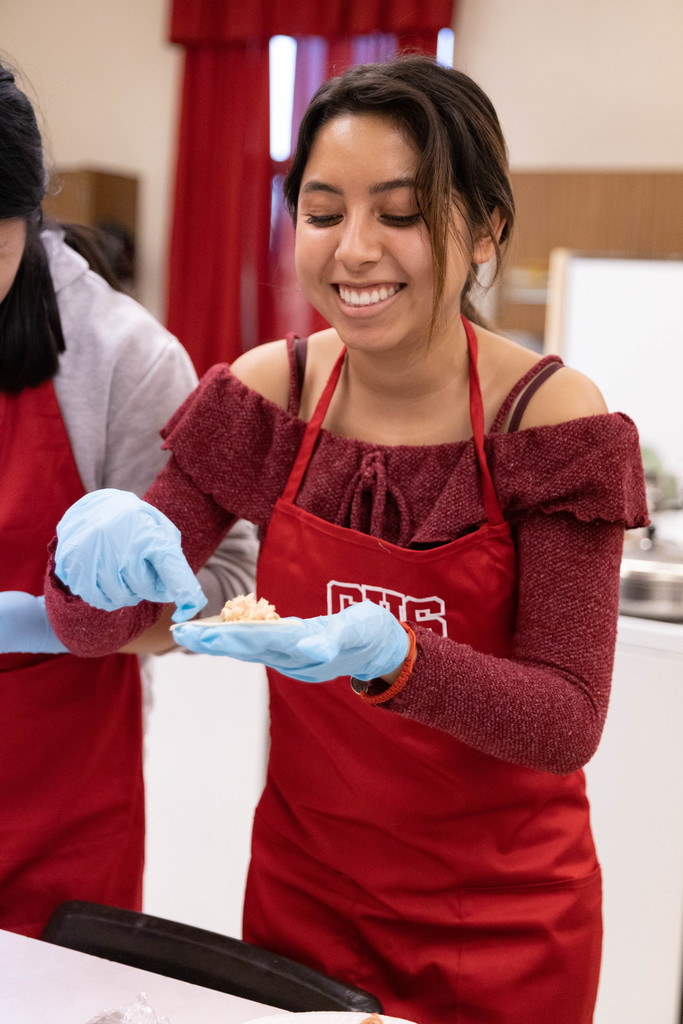 Female student in red apron holds potsticker in gloved hand
