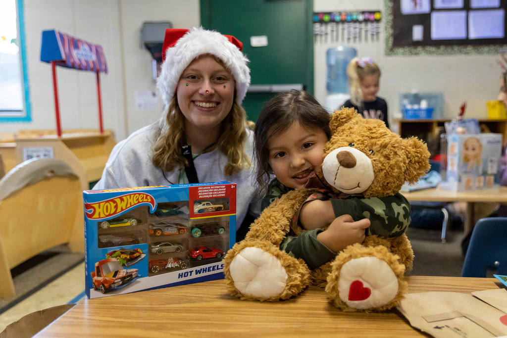 Female high school student in Santa hat sits next to elementary student holding teddy bear