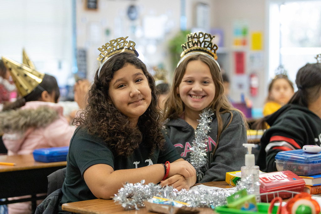 Two smiling young female students seated at desks and wearing "Happy New Year" tiaras