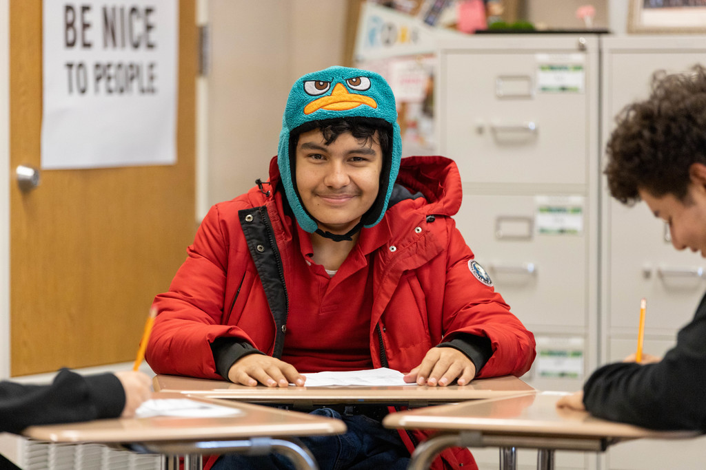 Male student in red jacket and winter hat seated at desk