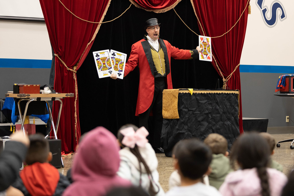 Magician in top hat and red coat holds out giant playing cards