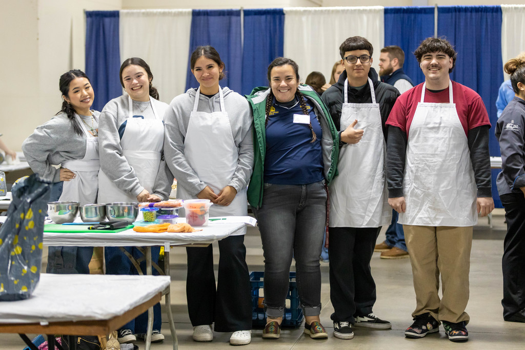 Students in white aprons stand near table holding containers of food they prepared