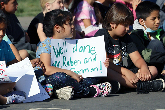 Students seated on blacktop hold celebratory signs