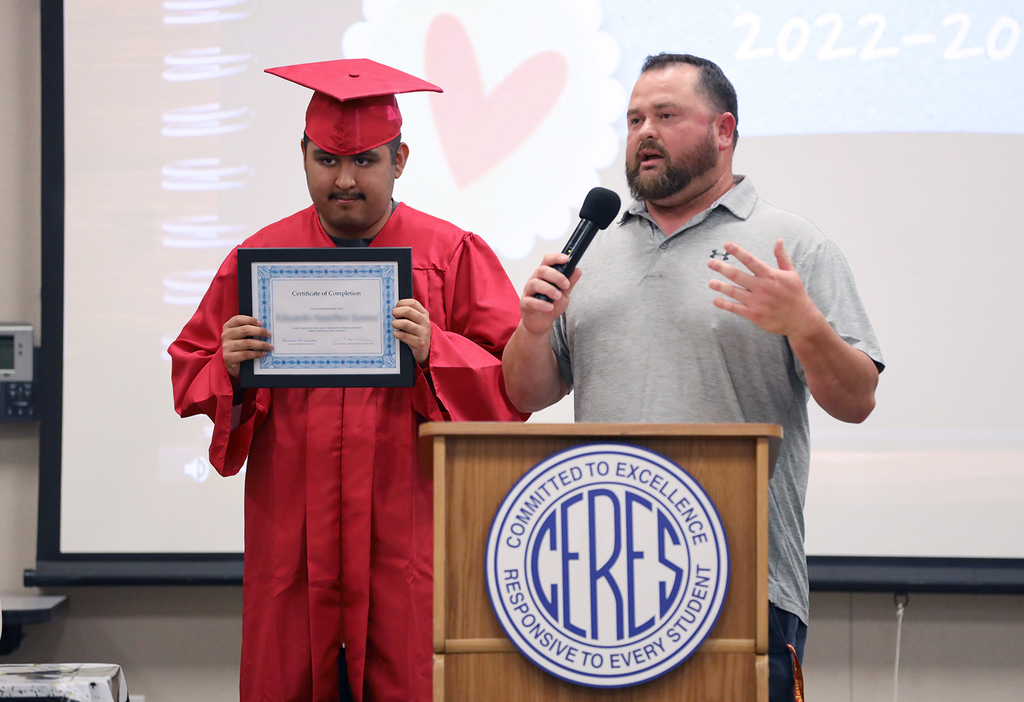 Student in red cap and gown holds certificate as teacher speaks into mic