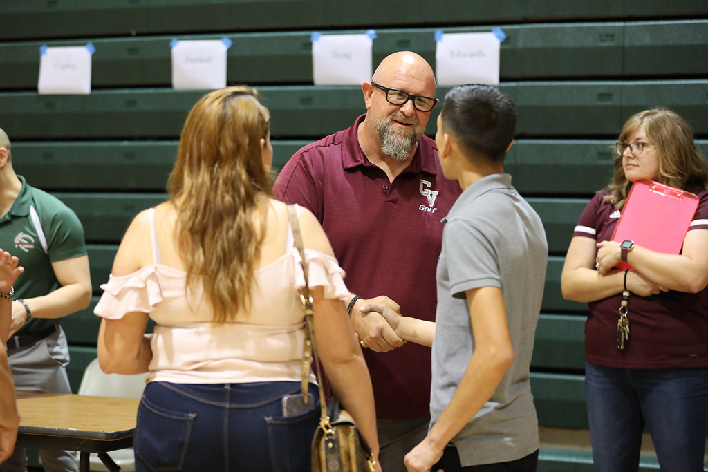 Teacher in school shirt shakes male student's hand while parent stands by